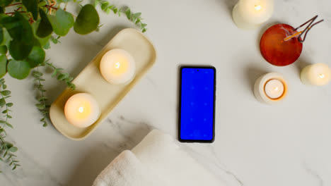 Overhead-View-Looking-Down-On-Still-Life-Of-Blue-Screen-Mobile-Phone-Lit-Candles-And-Incense-Stick-With-Green-Plant-And-Towels-As-Part-Of-Spa-Day-Decor